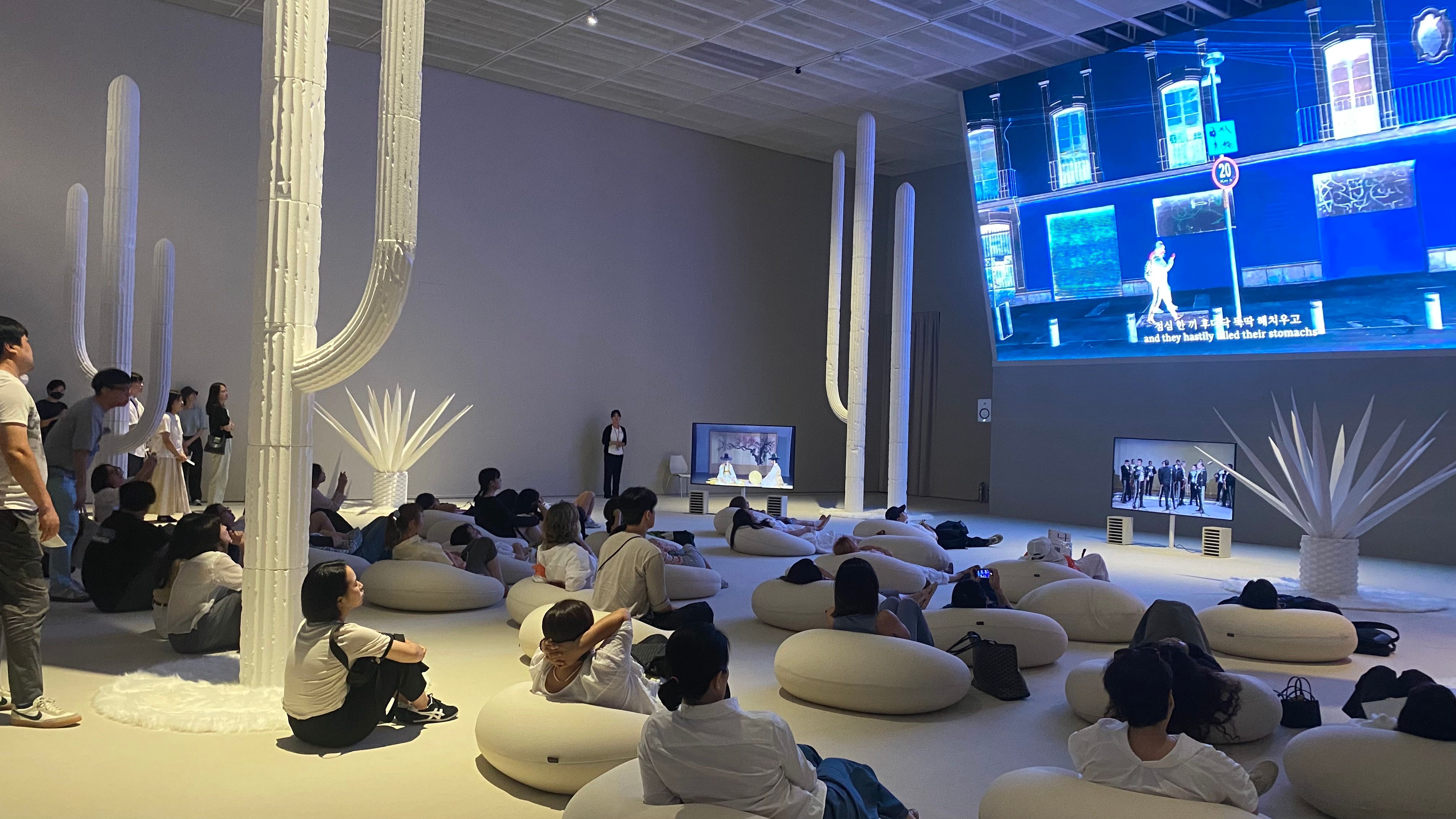 Visitors observe a TV in an art museum showing Jung Yeondoo's One Hundred Years of Travels, a mixed media installation with video, print, and sound.