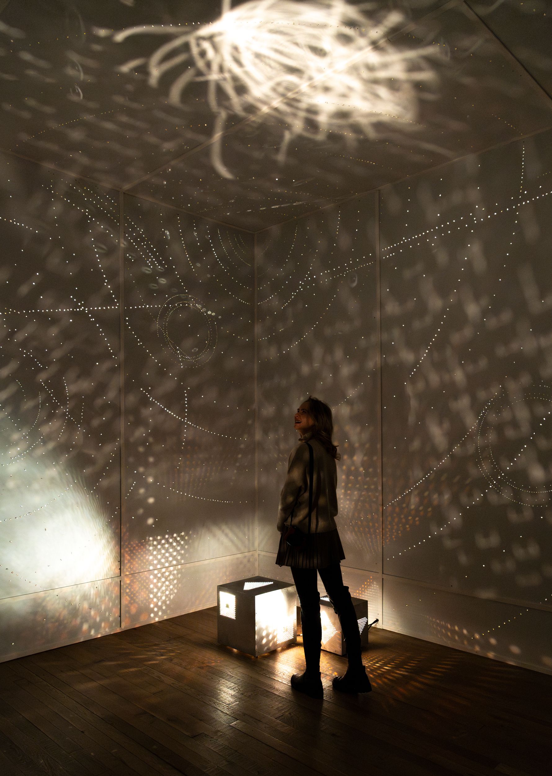 A woman stands inside a darkened room illuminated by intricate patterns of light and shadow cast onto the walls, ceiling, ...