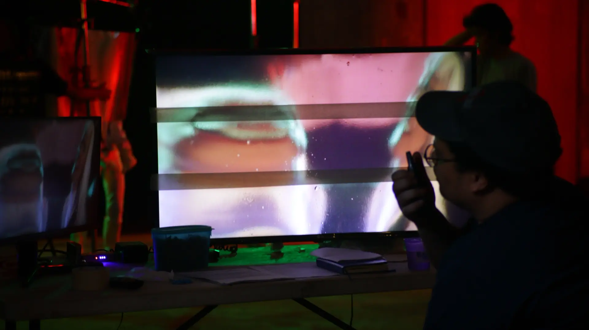 An individual is seen concentrated at a monitor, speaking into a handheld device. They are situated in a professional setting, suggesting a behind-the-scenes look at a technical process.