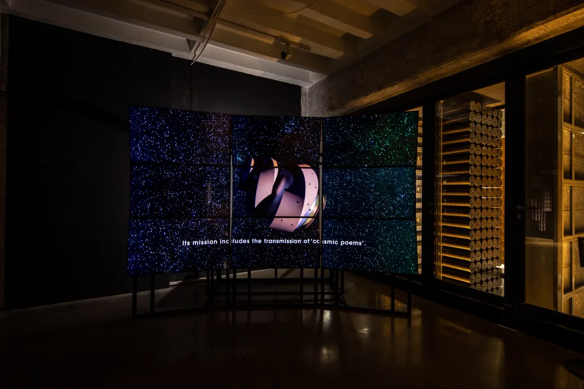 A dark gallery hall displays a cosmic sky image across nine monitors, forming one connected picture. An undefined iron shape is central to the scene, and the subtitle reads "Its mission includes the transmission of cosmic poems".