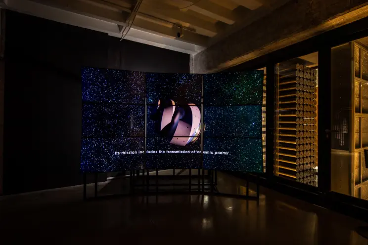 A dark gallery hall displays a cosmic sky image across nine monitors, forming one connected picture. An undefined iron sha...