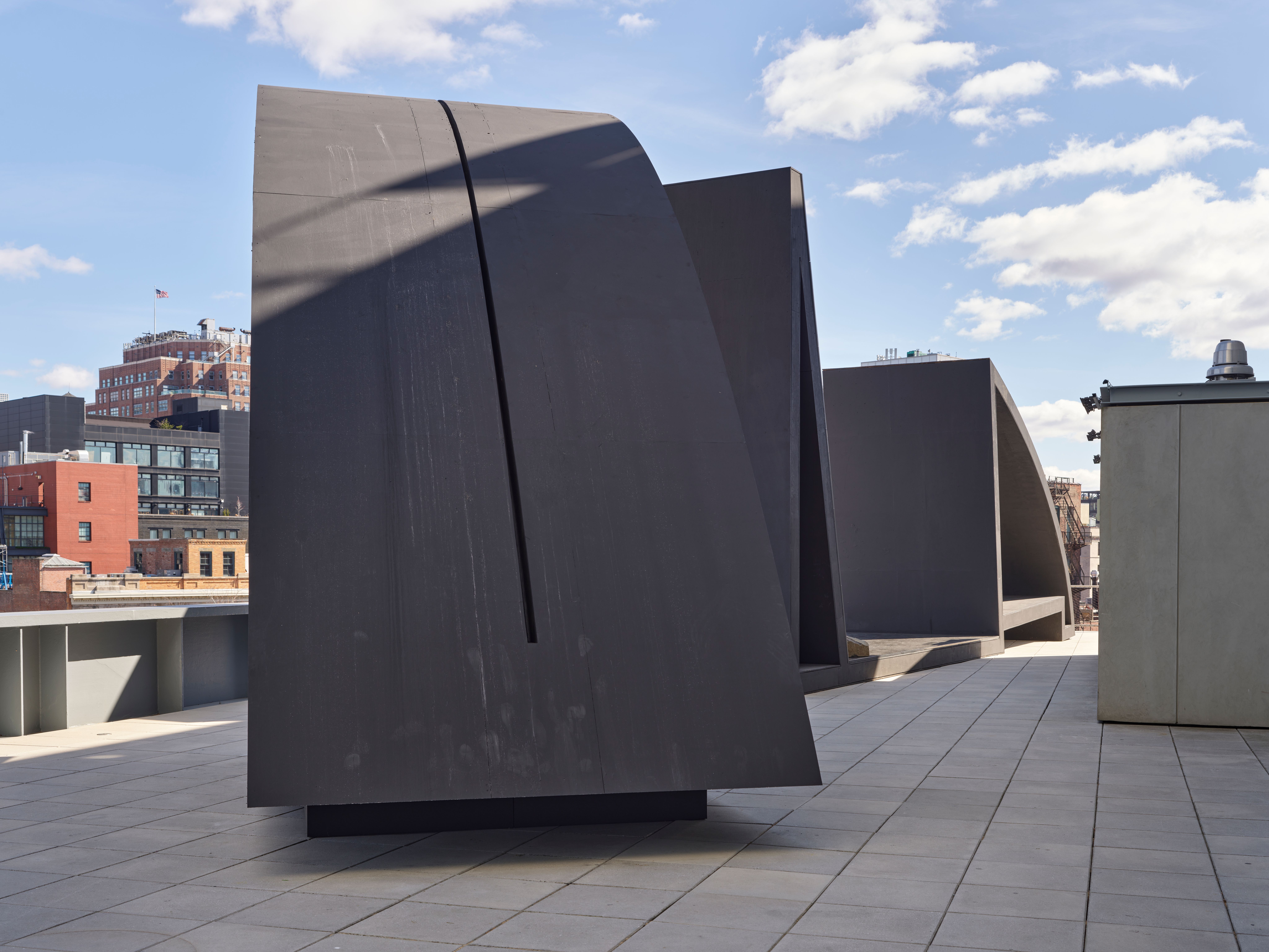 Large, dark modern sculptures on a rooftop terrace with city buildings and a blue sky in the background.