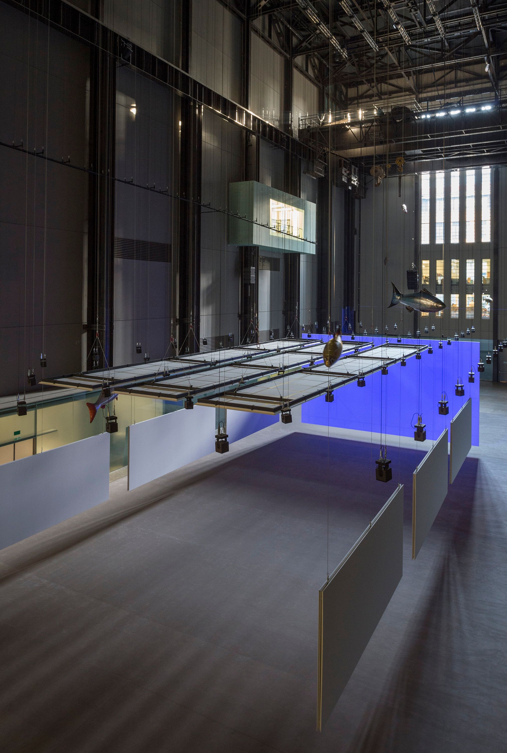An art installation is displayed in a spacious gallery hall, featuring multiple fish sculptures hanging from the ceiling.