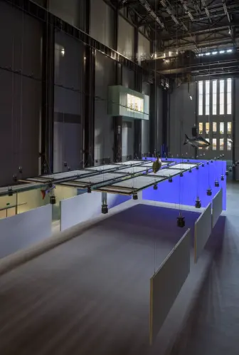 An art installation is displayed in a spacious gallery hall, featuring multiple fish sculptures hanging from the ceiling.