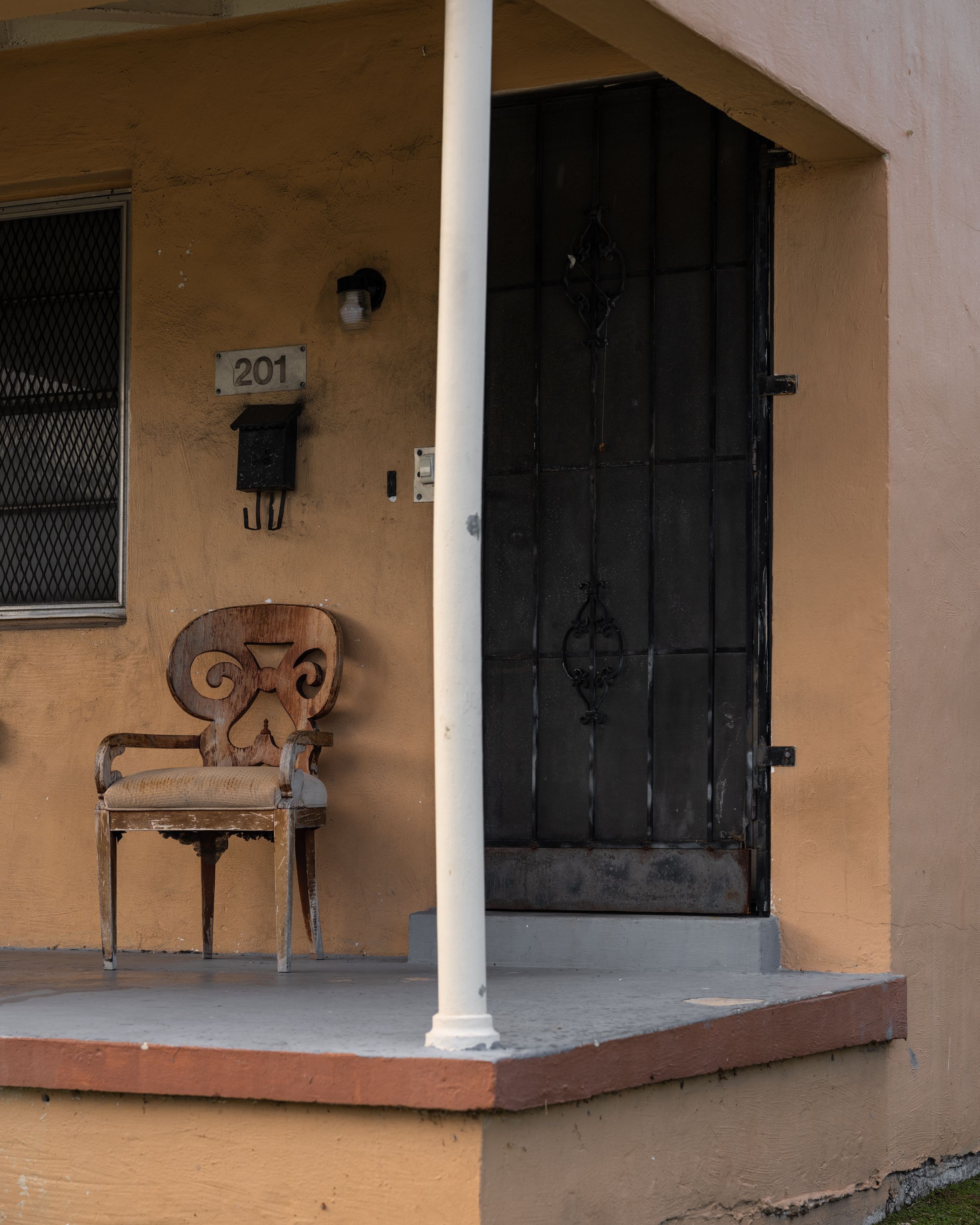 A lone wooden chair sits on the front porch of a yellow house with the number 201, featuring a black metal door and a mailbox.