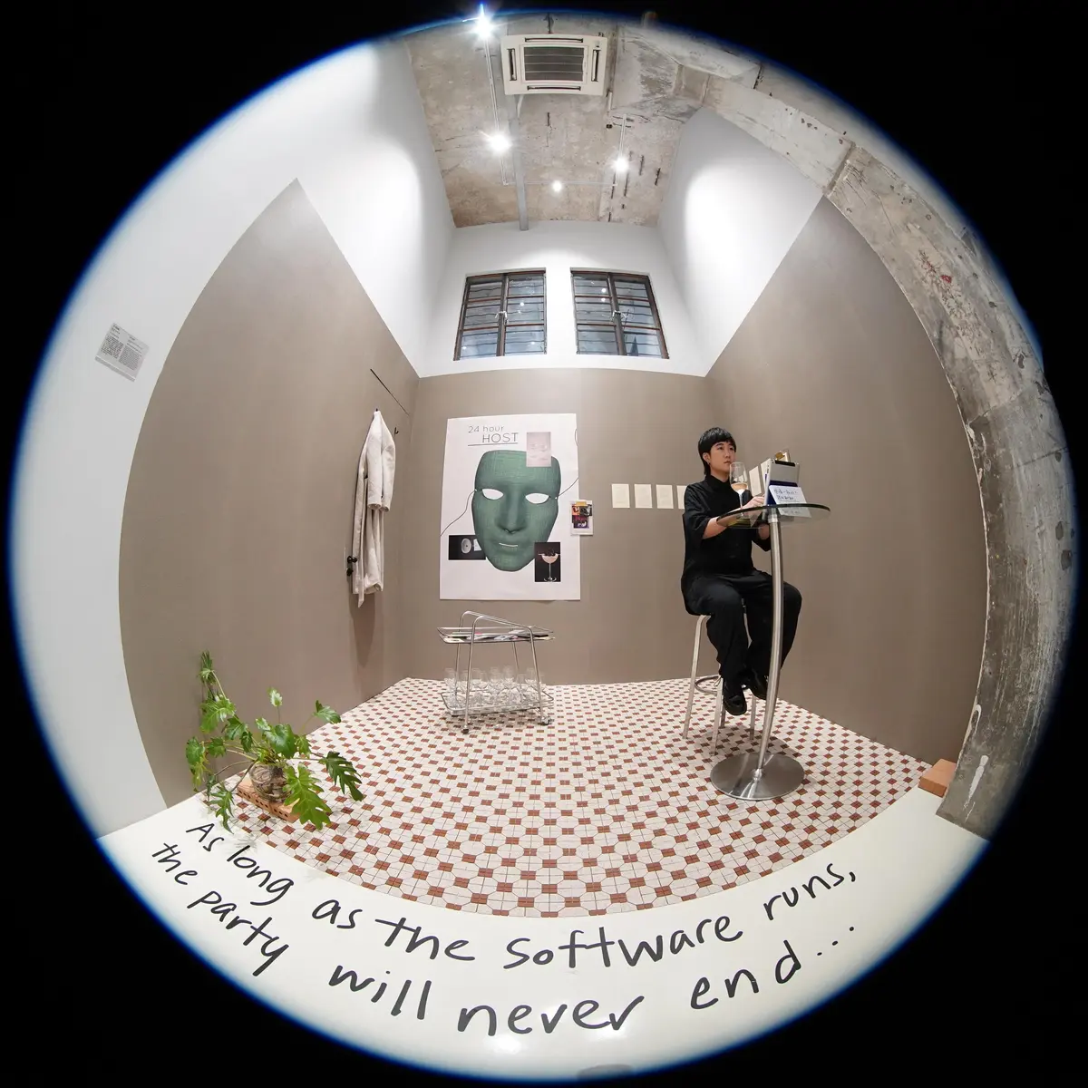 A person sitting at a high top table in a room captured through a fisheye lens. The room contains a poster of a green face mask, a hanging white coat, and a note on the floor that reads, "As long as the software runs the party will never end."