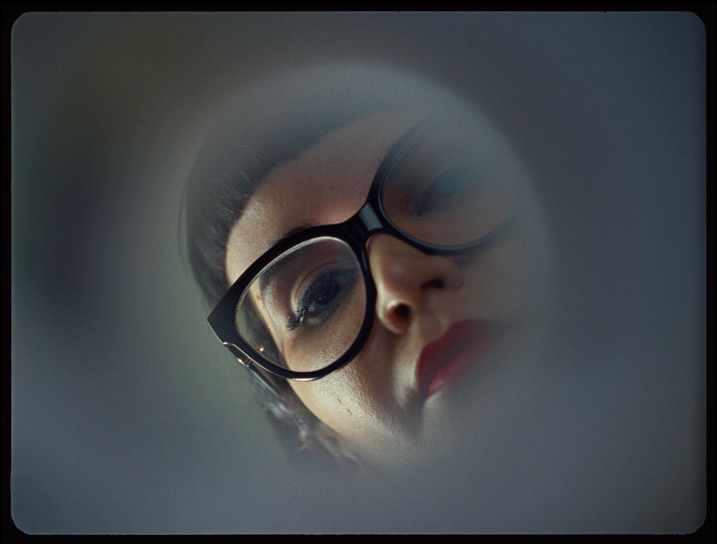 A close-up of a woman's face with glasses and red lipstick, partially obscured through a blurred surface.