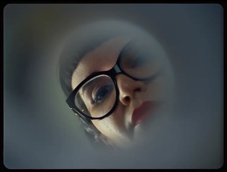 A close-up of a woman's face with glasses and red lipstick, partially obscured through a blurred surface.