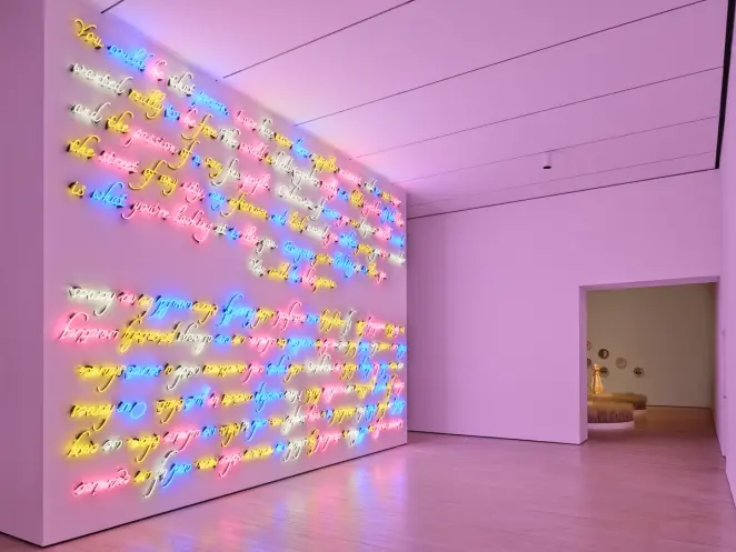 Large wall of multicolored neon cursive text glowing in a dim gallery, viewed from an angle with doorway beyond.