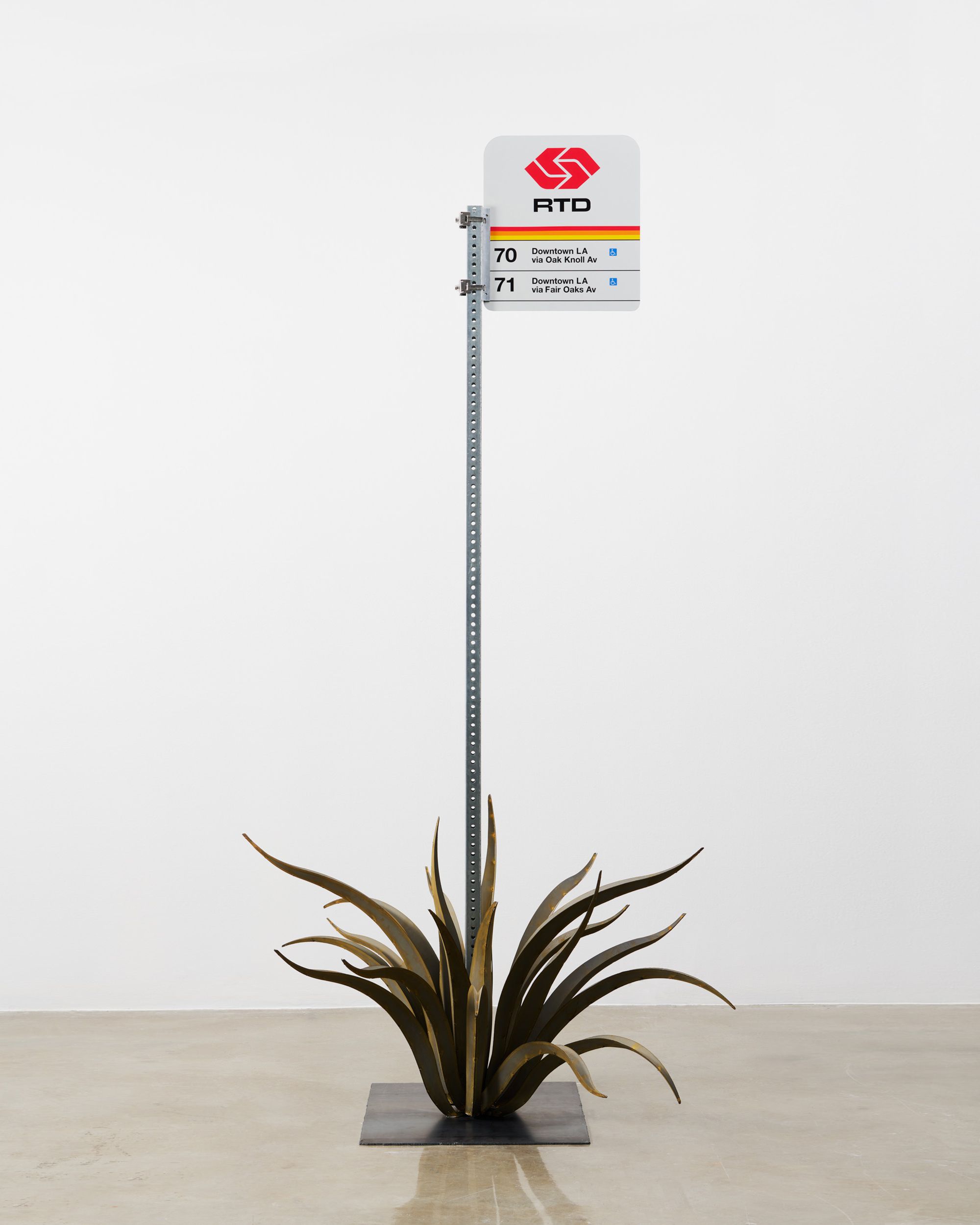 A minimalist sculpture of a Los Angeles bus stop sign with agave-like metal leaves at its base.