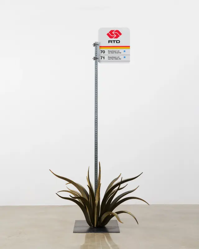 A minimalist sculpture of a Los Angeles bus stop sign with agave-like metal leaves at its base.