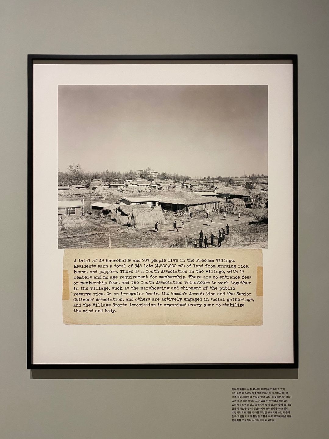 A vintage photograph of a town, depicted in black-and-white tones, mounted in a frame on the wall of an art gallery. The p...