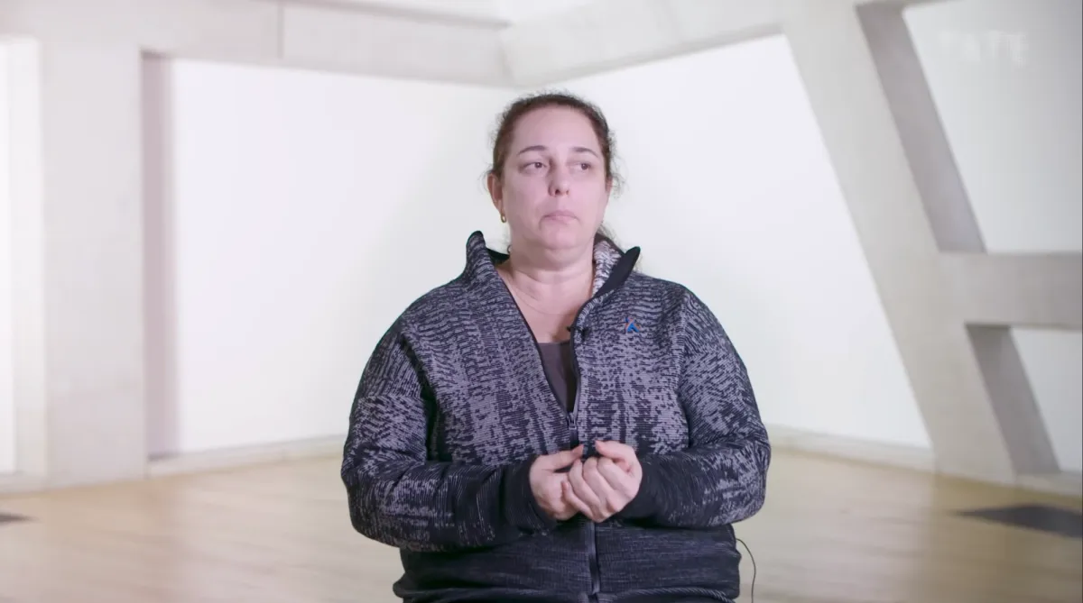 A video still of Tania Bruguera, an artist captured mid-gesture as they address the camera in dialogue.