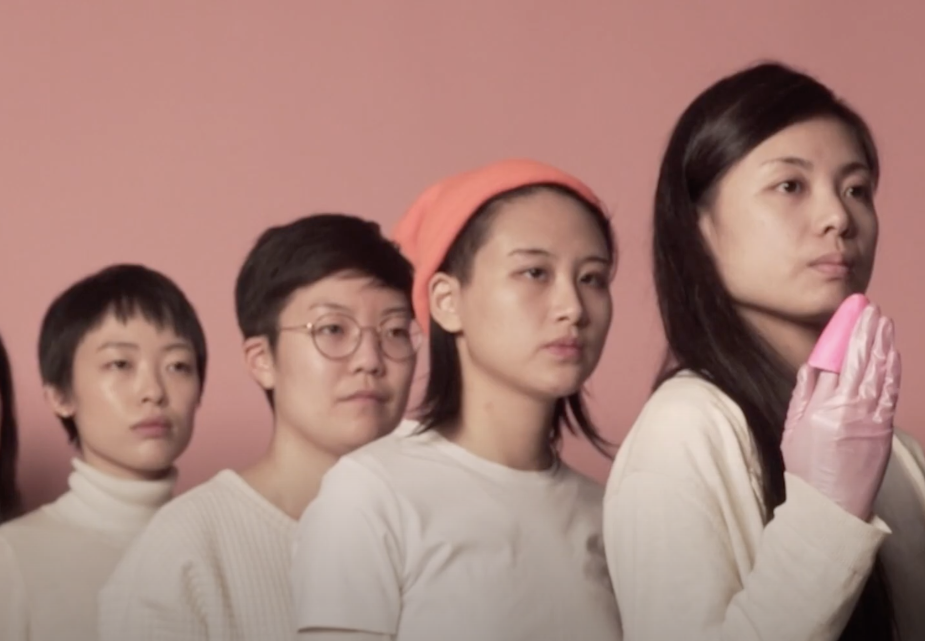 A group of individuals in white outfits and pink gloves form a line on a pink background. The person at the front extends ...