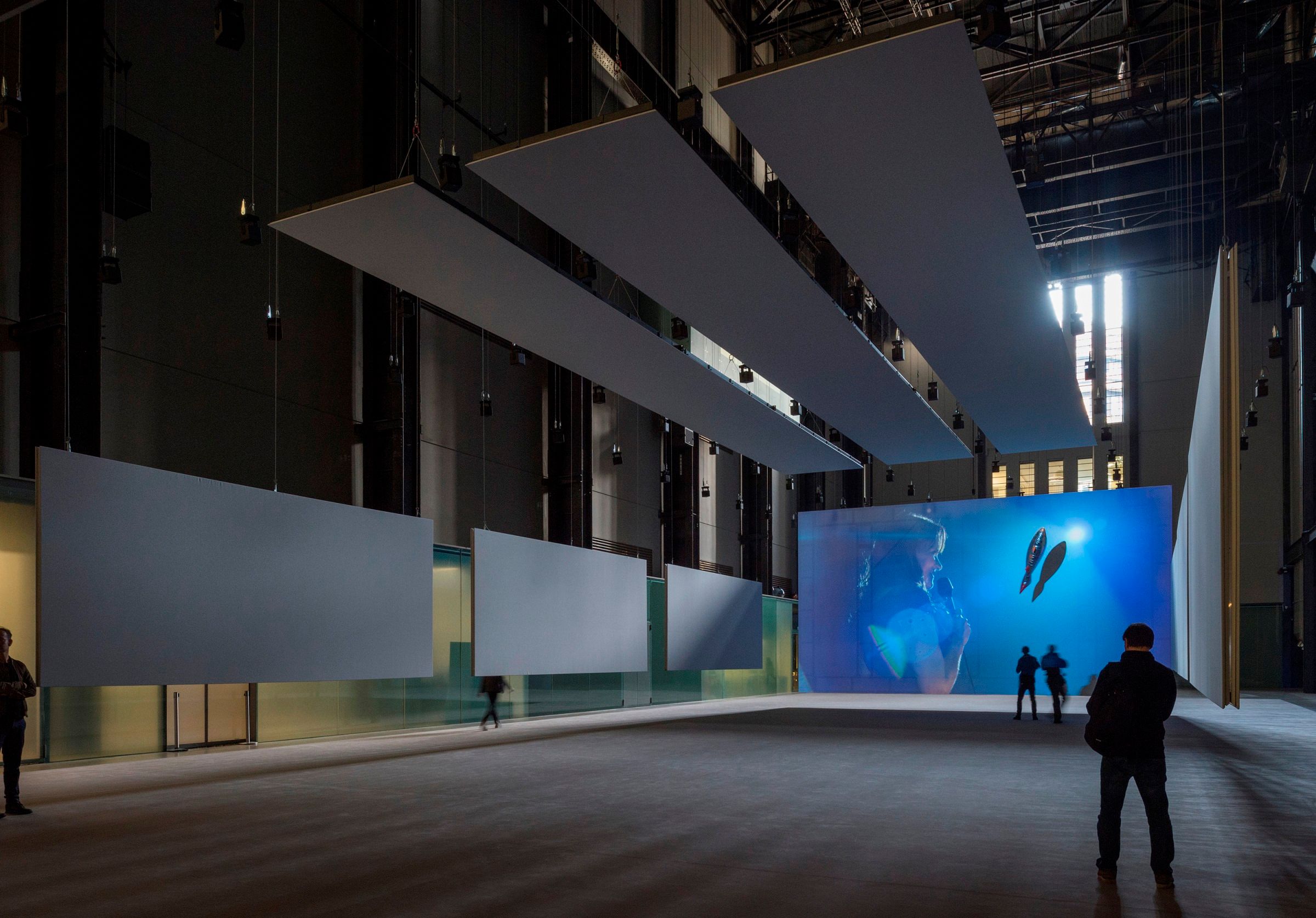 A spacious gallery hall in Tate Modern featuring white panel ornaments on the sides and ceiling. A person holds a micropho...