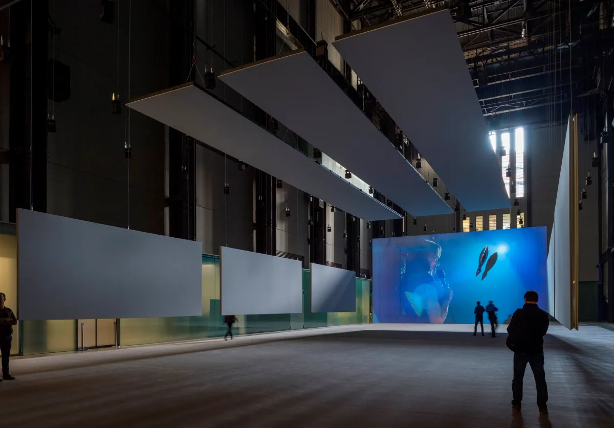 A spacious gallery hall in Tate Modern featuring white panel ornaments on the sides and ceiling. A person holds a microphone on the left near a wide screen displaying an underwater photo. Few visitors walk about.