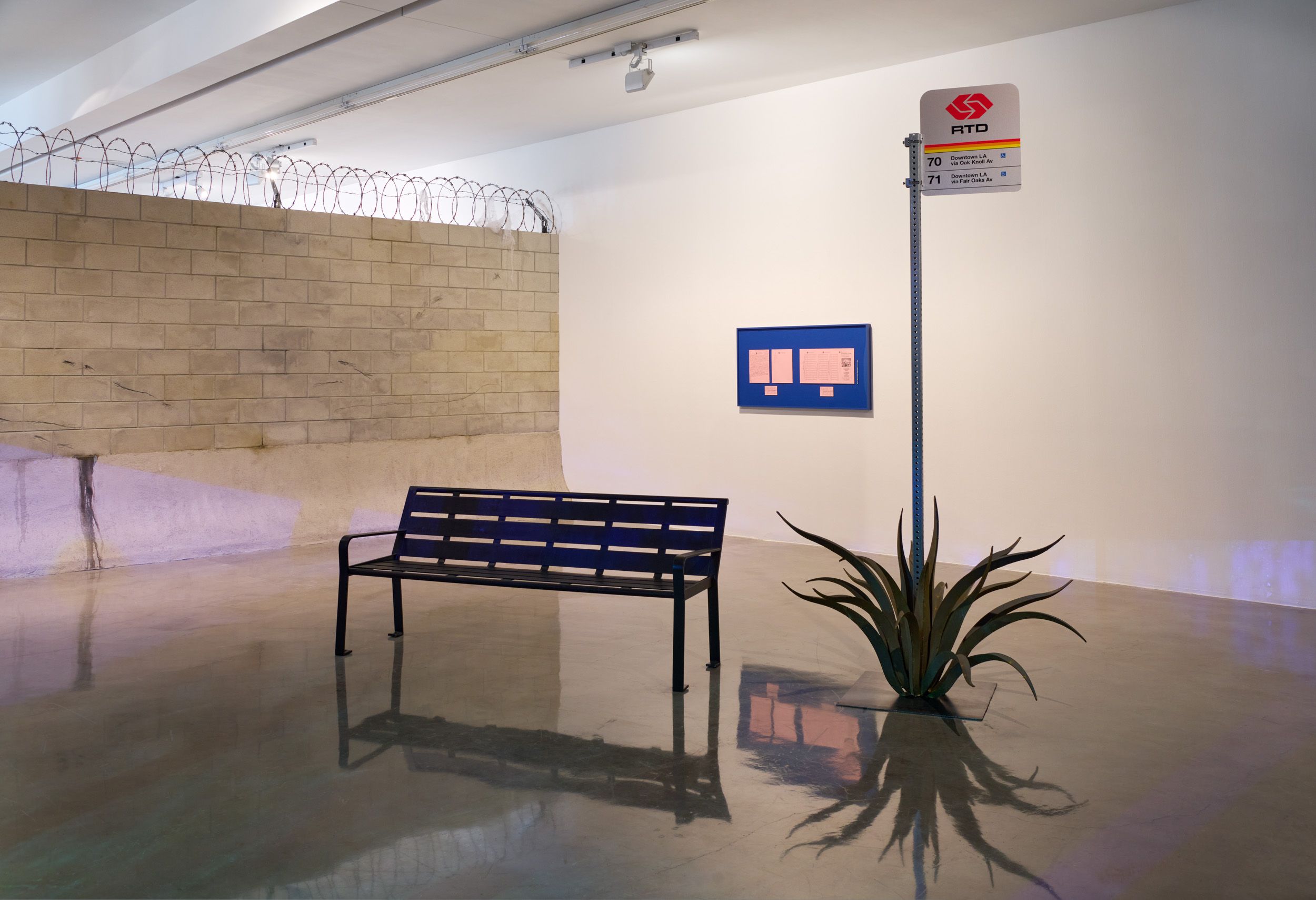 An installation of a bus stop sign with a bench and a barbed-wire-topped wall, creating an urban streetscape in a gallery.