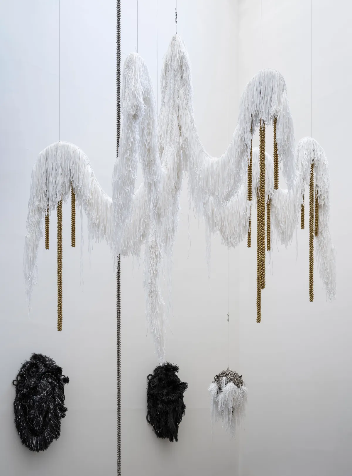 An art installation featuring a large, white, furry chandelier-like structure suspended from the ceiling. Adorning the walls are two dark, furry forms, with a smaller white, furry spherical object also hanging from above.
