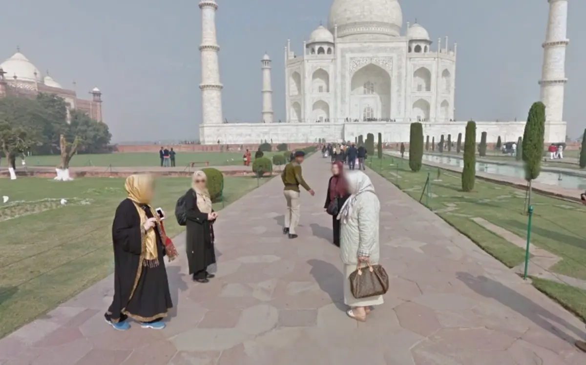 A pathway outdoors with passersby, leading the eye towards the Taj Mahal in the background under a clear blue sky.