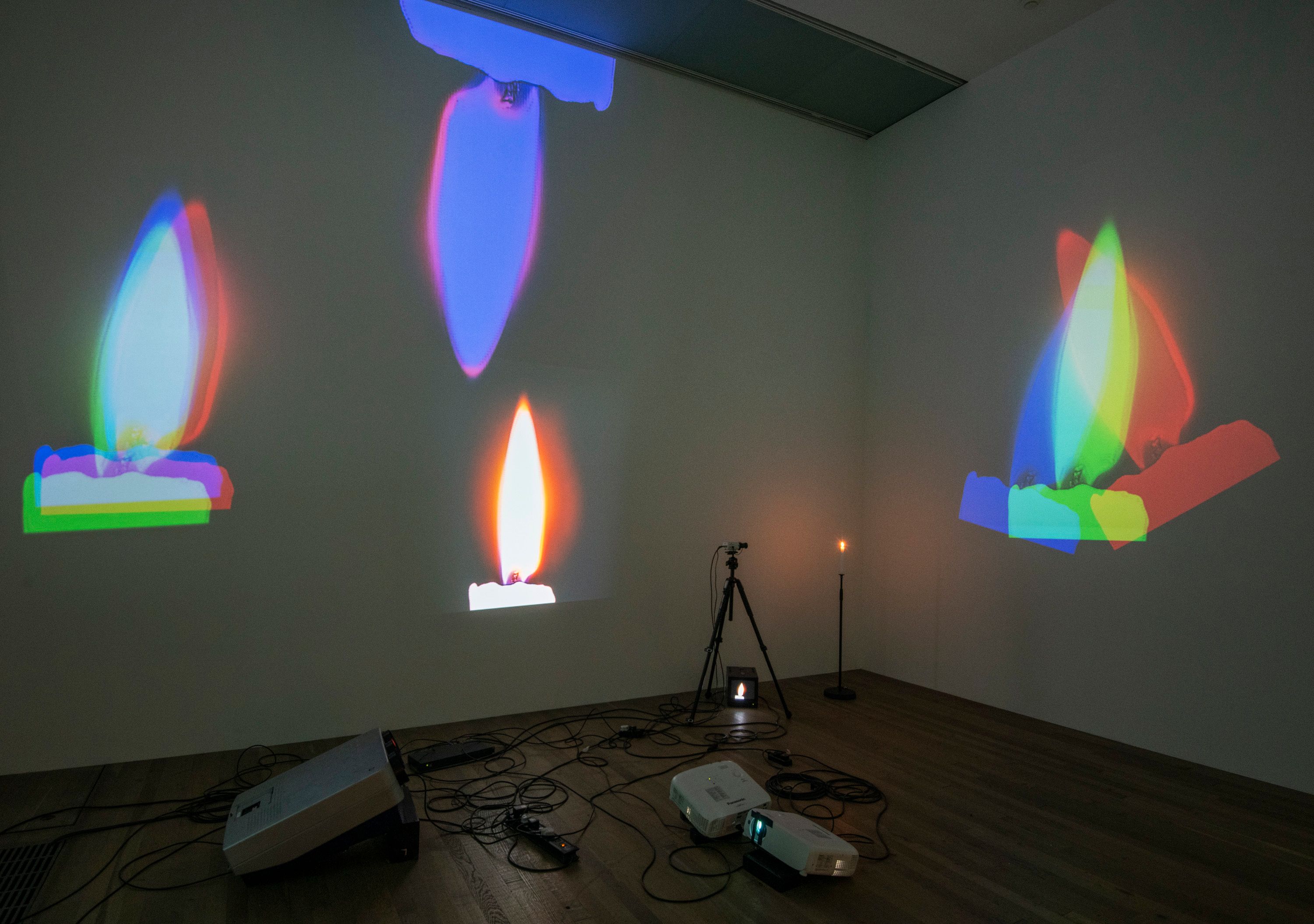 A projection of colorful flame images on a white wall in a room, with visible projection equipment and wires on a wooden f...