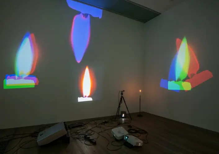 A projection of colorful flame images on a white wall in a room, with visible projection equipment and wires on a wooden f...