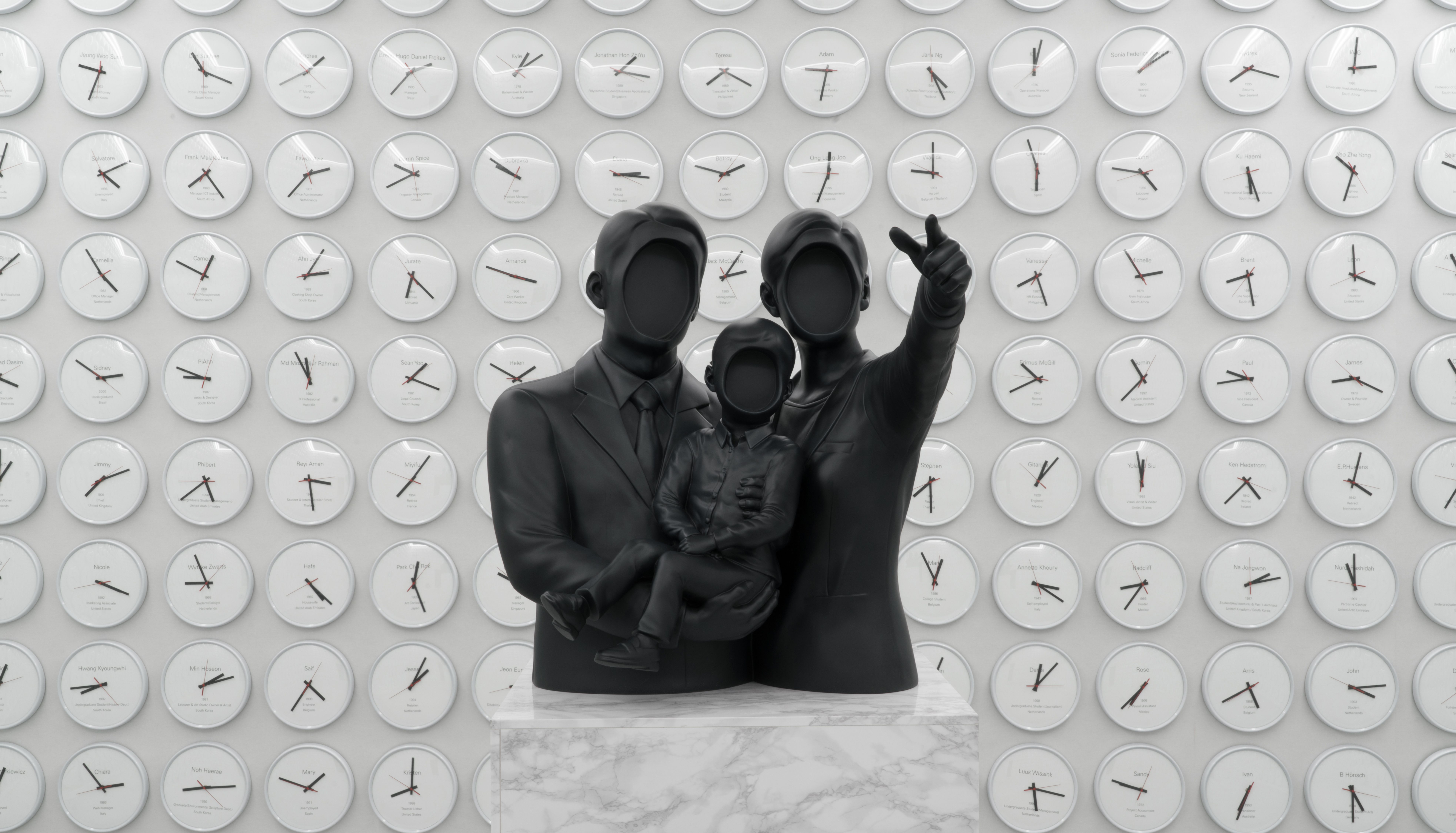 A minimalist black sculpture of a faceless family, composed of two adults and a child, set against a white wall adorned wi...