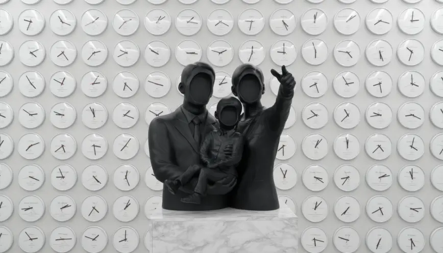 A minimalist black sculpture of a faceless family, composed of two adults and a child, set against a white wall adorned wi...