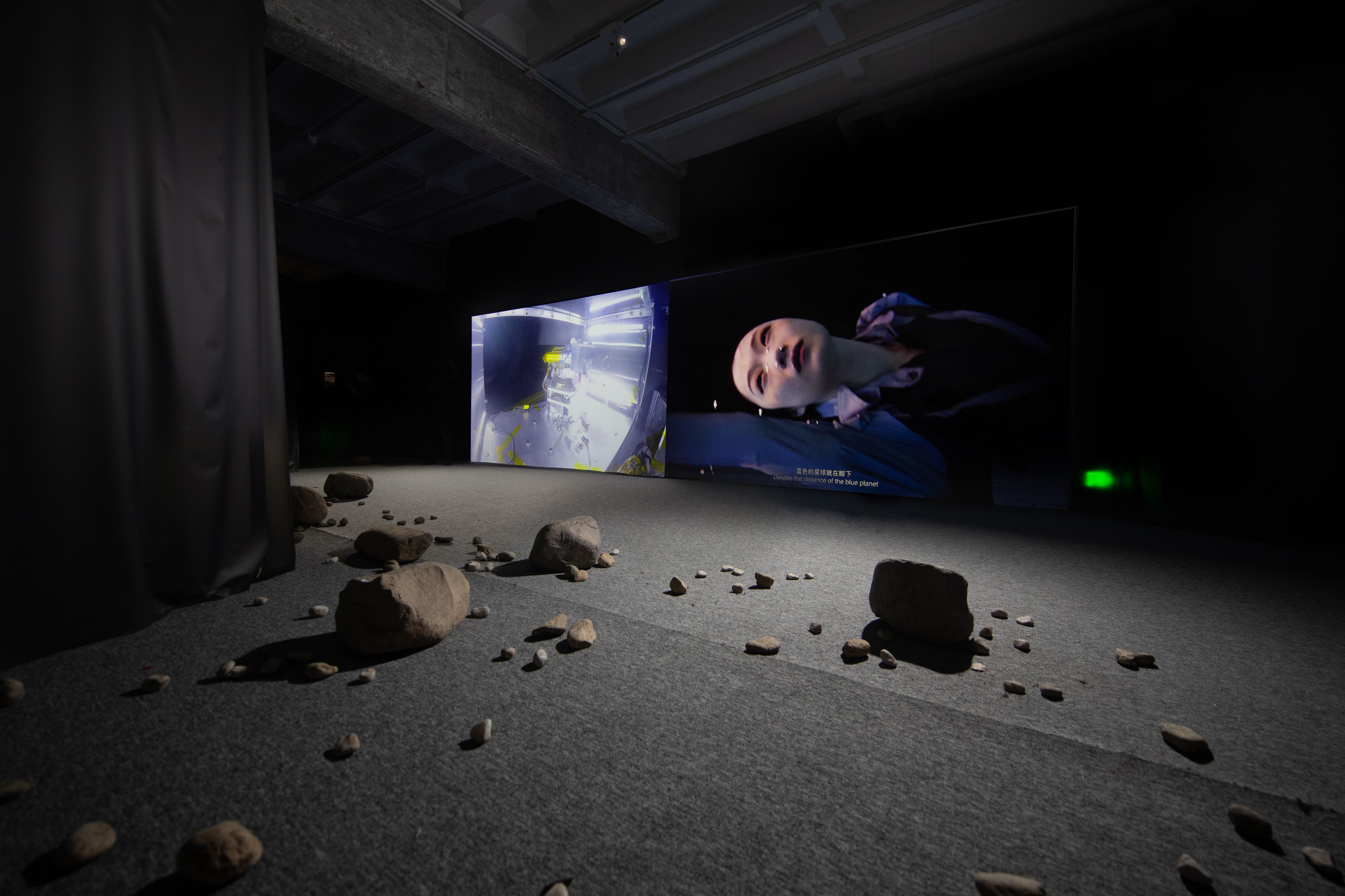 A dimly lit exhibition room displaying various large and small rocks scattered across the floor. In the room, a screen dis...