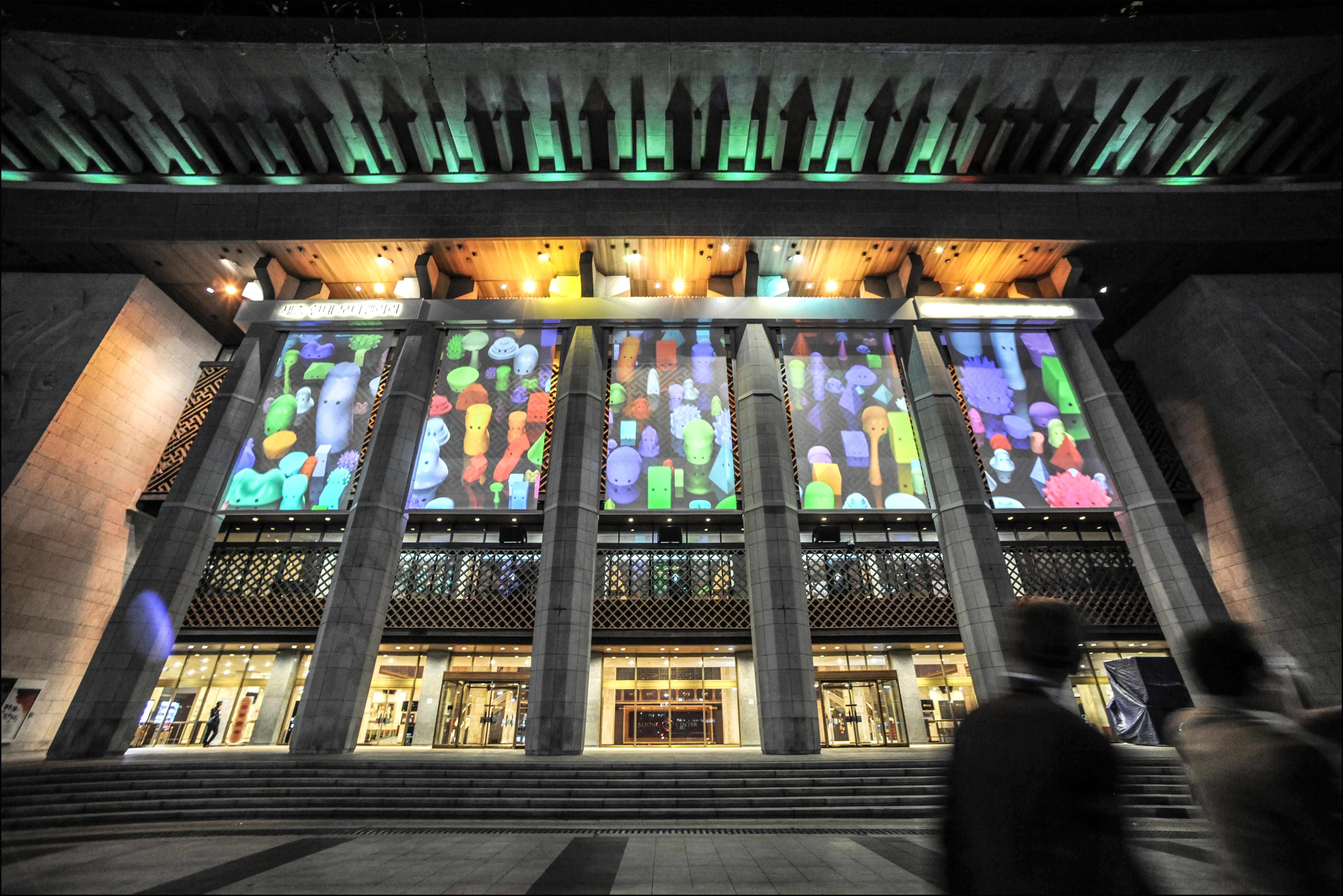 A large building's façade showcases lighted pictures on its face. The structure has steps leading up to it, punctuated by ...