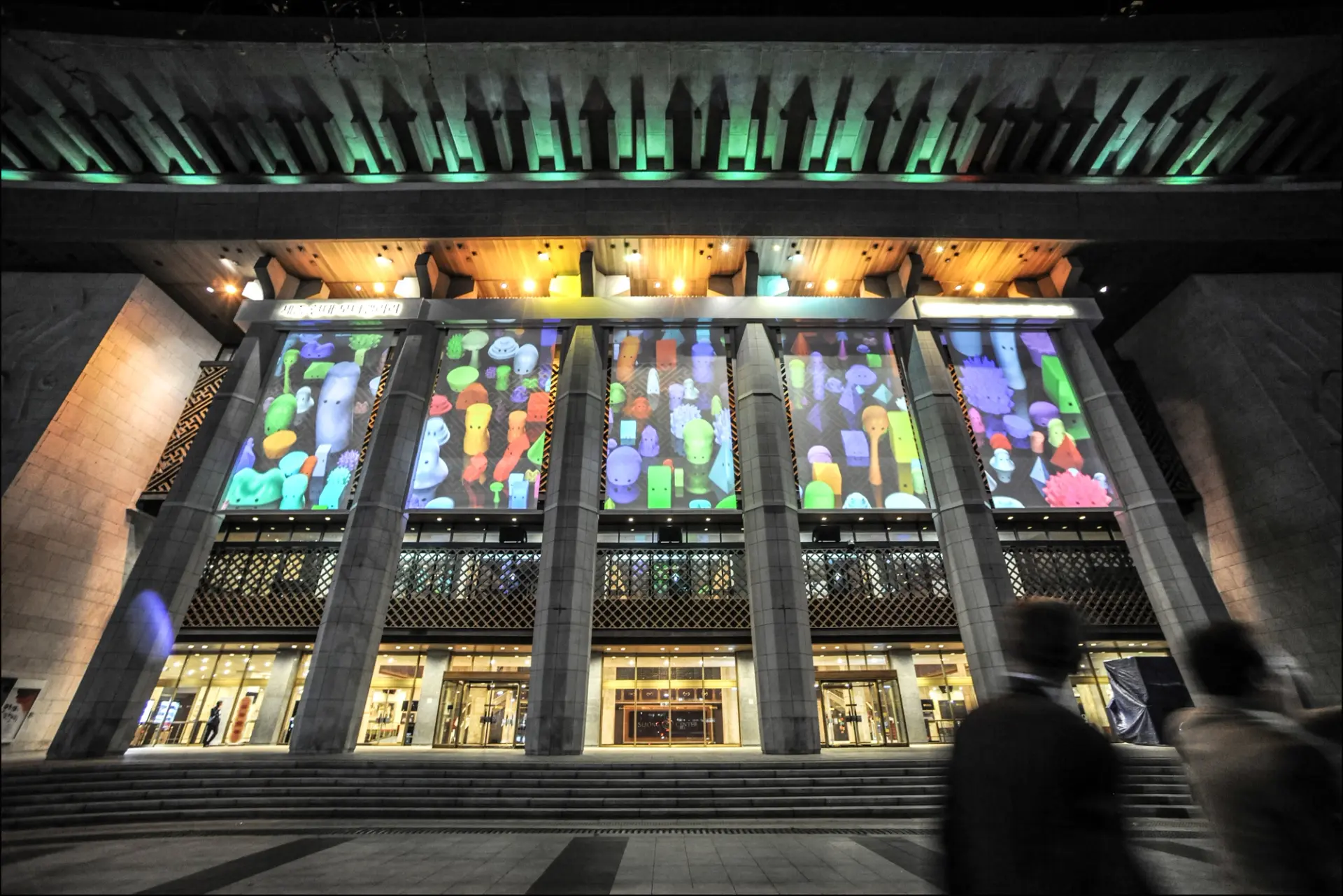 A large building's façade showcases lighted pictures on its face. The structure has steps leading up to it, punctuated by ...