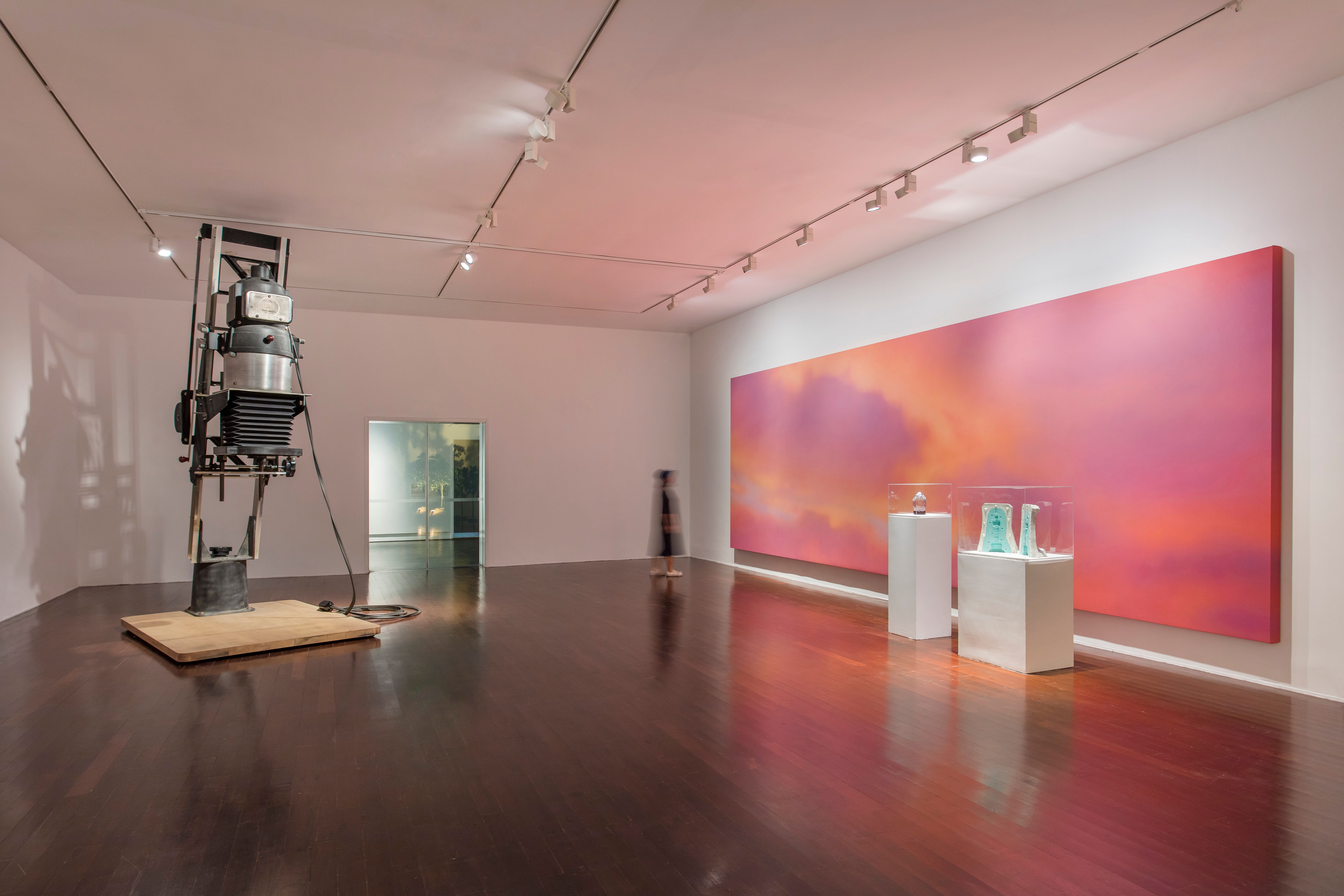 A gallery room showcasing a wide canvas with pink art on the wall. In front of it, two other art pieces are exhibited. A b...