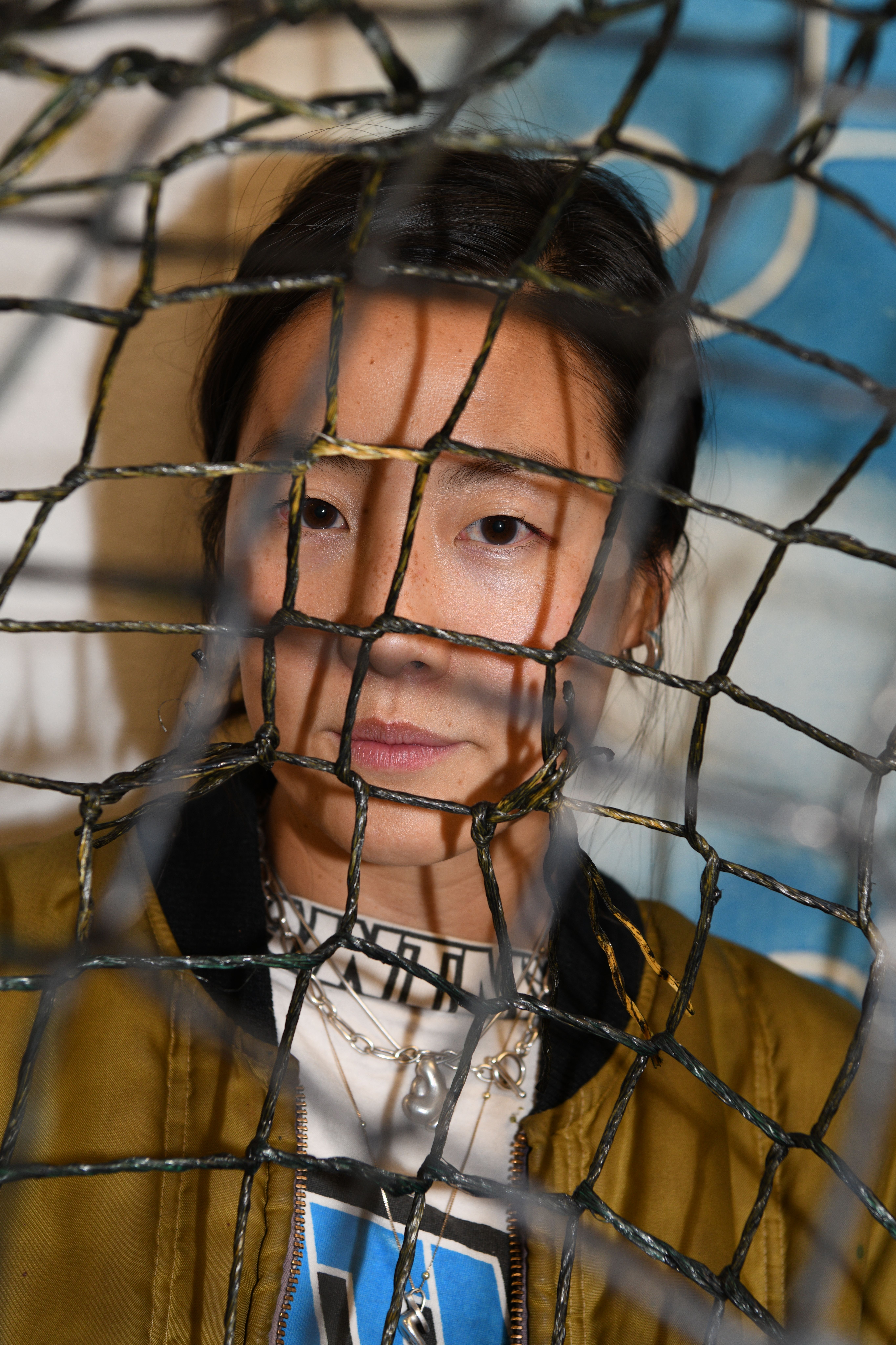 Portrait of an artist peering thoughtfully through a wire mesh, surrounded by elements of her creative workspace.
