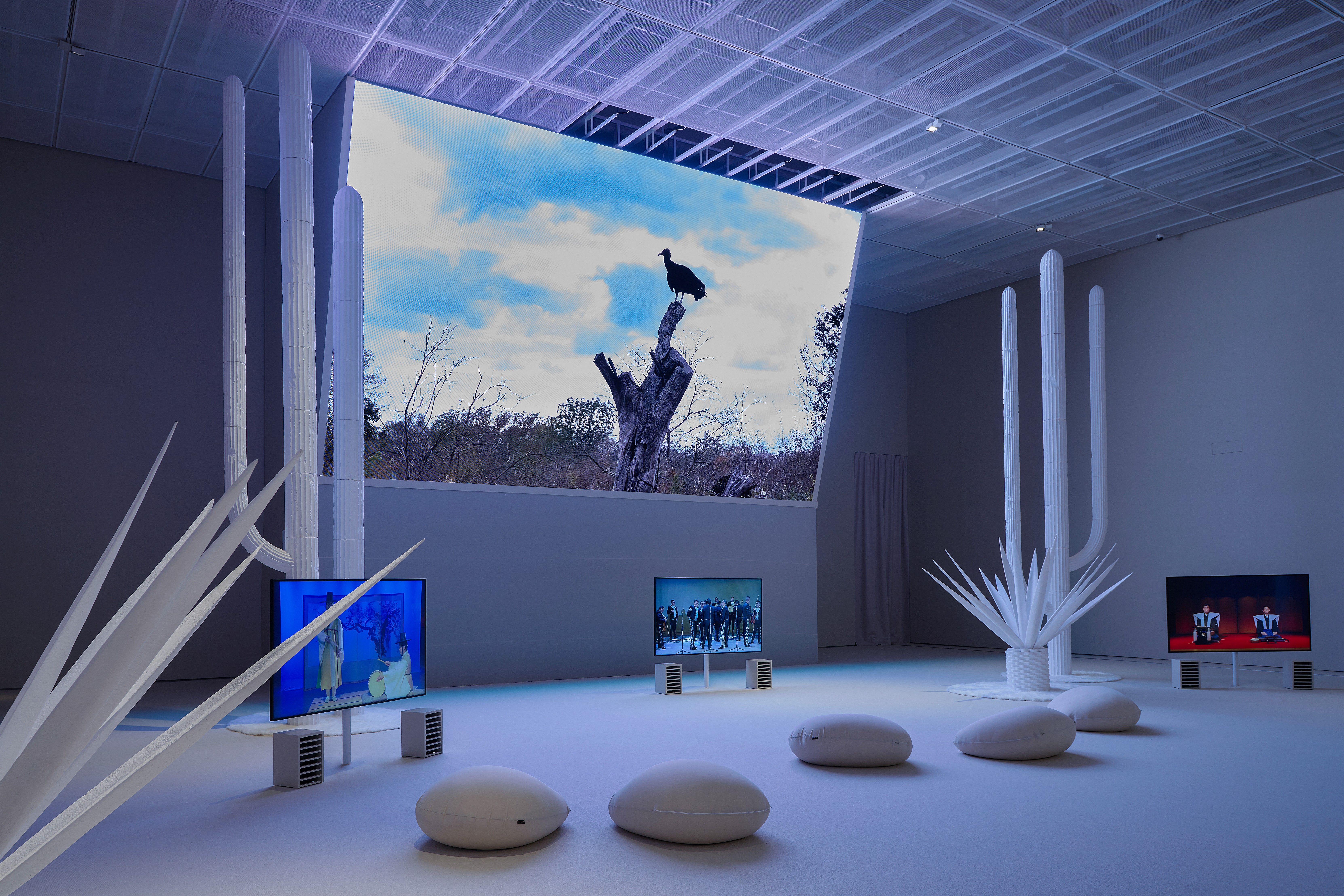 Contemporary art exhibition with white sculptures and monitors displaying vivid images in a blue-lit gallery space.
