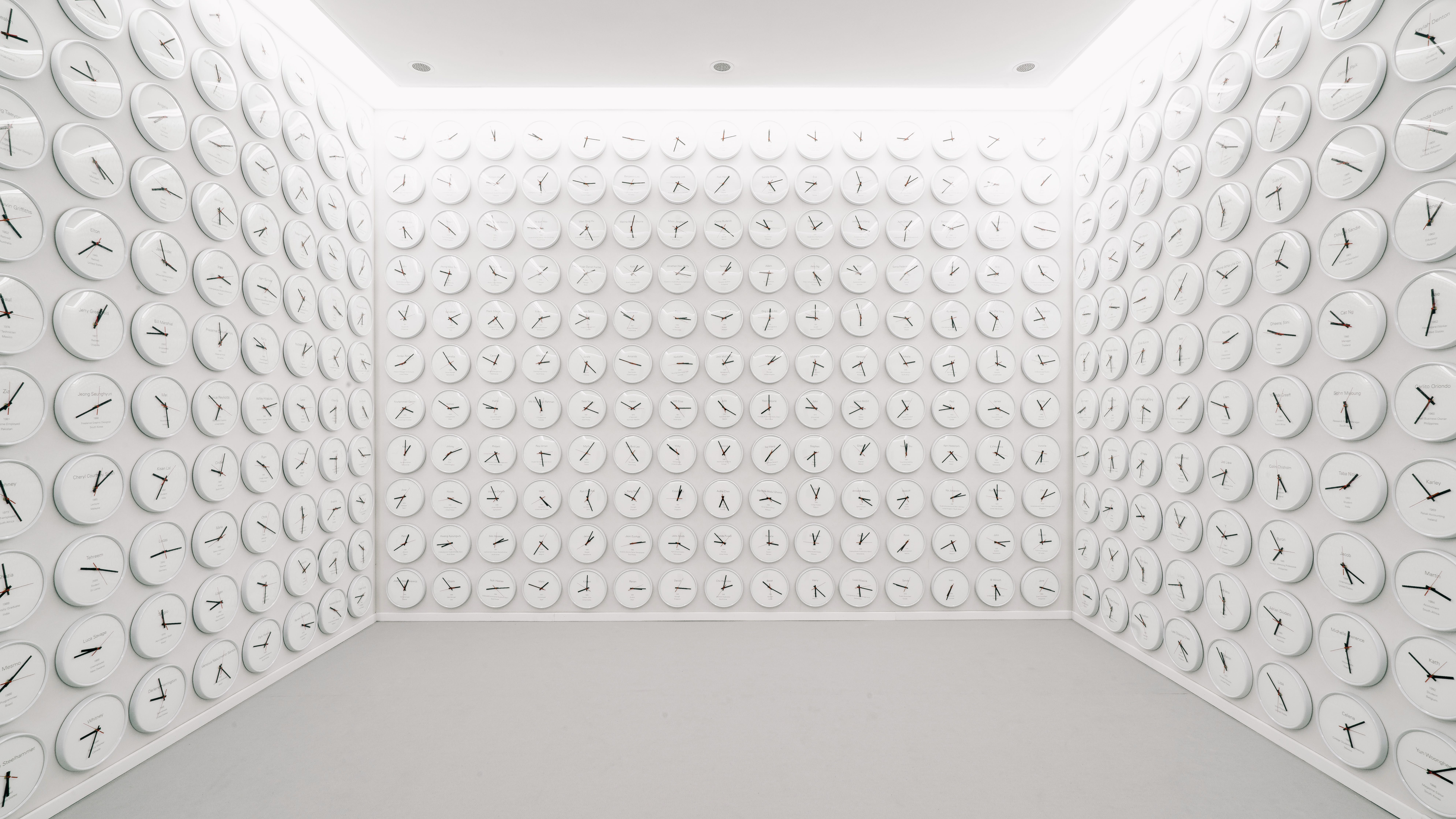 A room with walls entirely adorned with round white clocks. Each clock displays a different time, indicative of various gl...