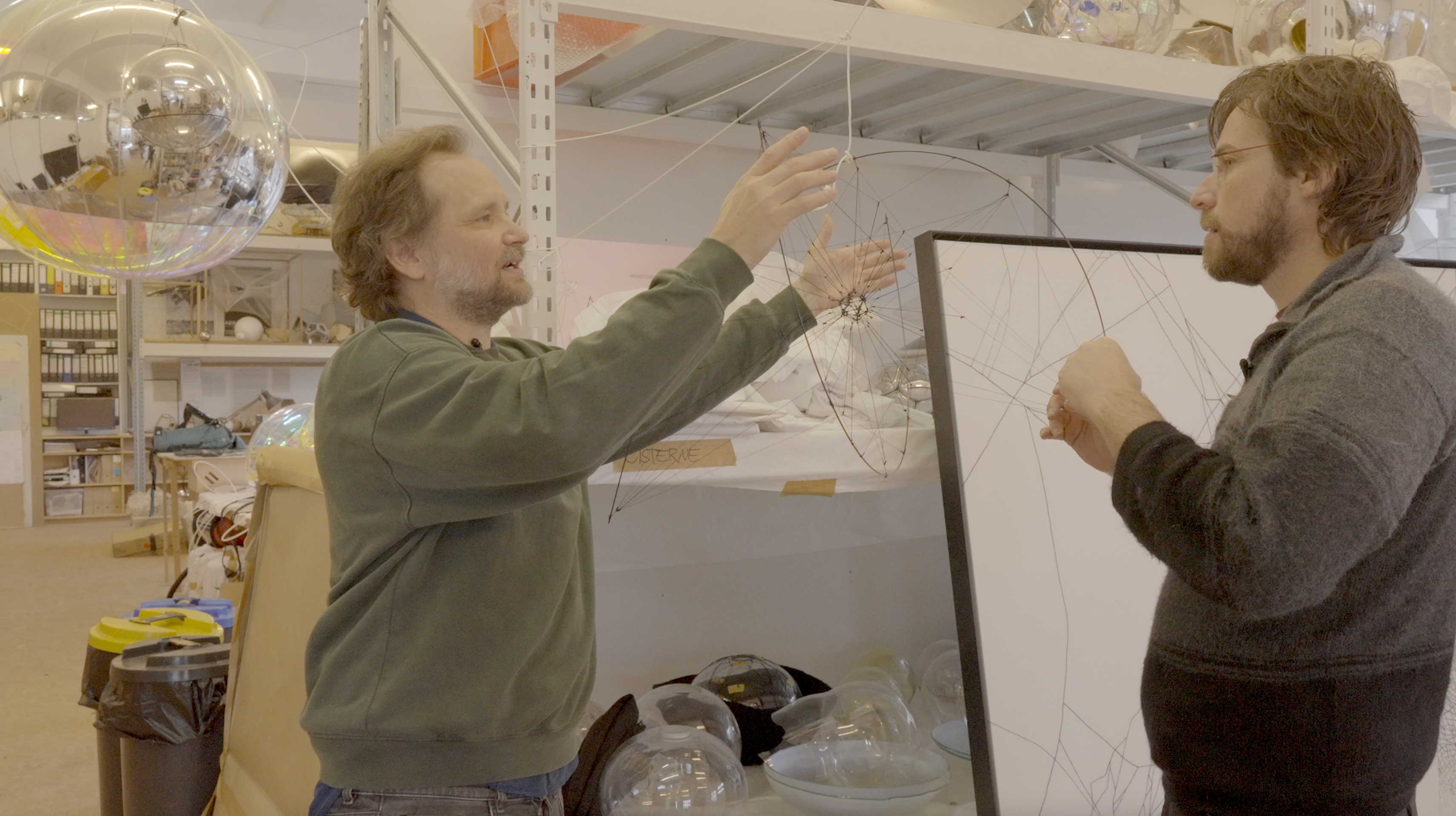 Two men discussing a web-like sculpture in a workshop filled with various creative materials.