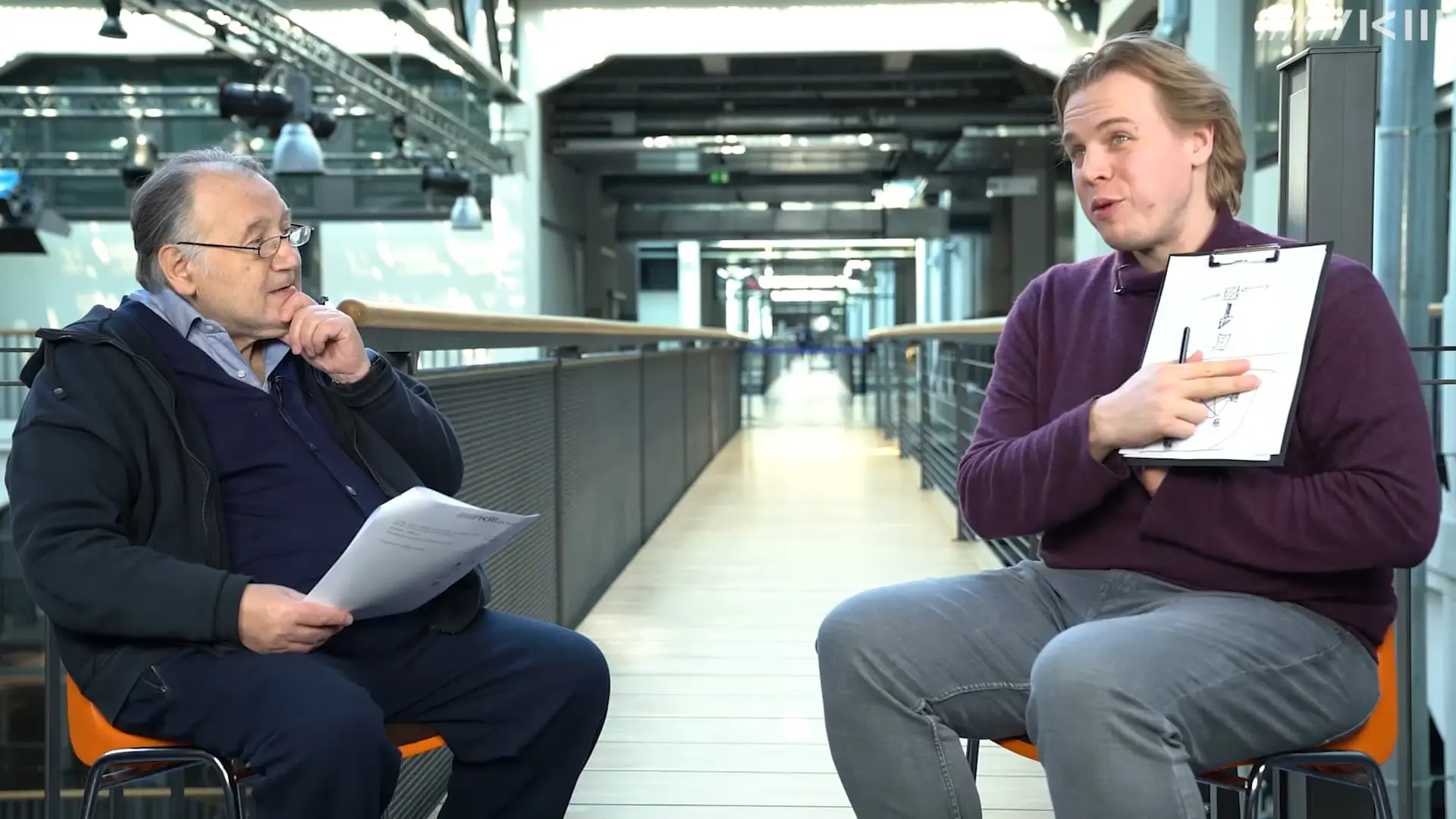 Two men in conversation, one holding papers, in a modern indoor space.