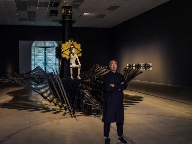 A person observes an intricate installation, topped by a human figure and featuring yellow hexagons.