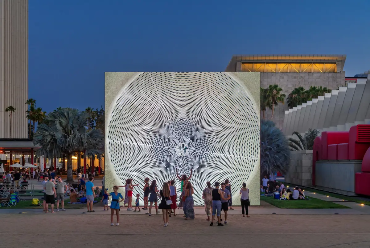 A group of individuals gather around an outdoor art installation featuring intricate circular patterns, observing it in the soft glow of evening. Palm trees rise in the background, silhouetted against the twilight sky.