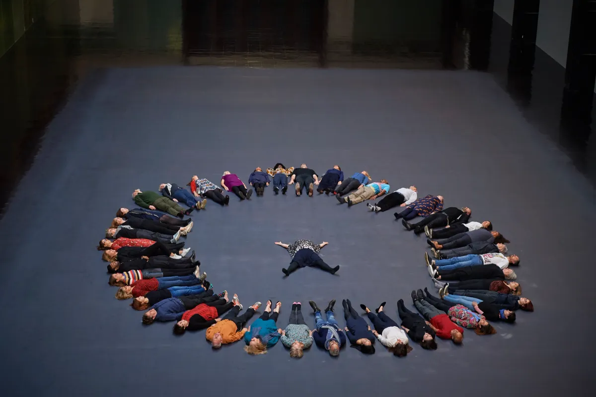 People form a circle while lying on a dark blue floor, with one individual spread out at the center.