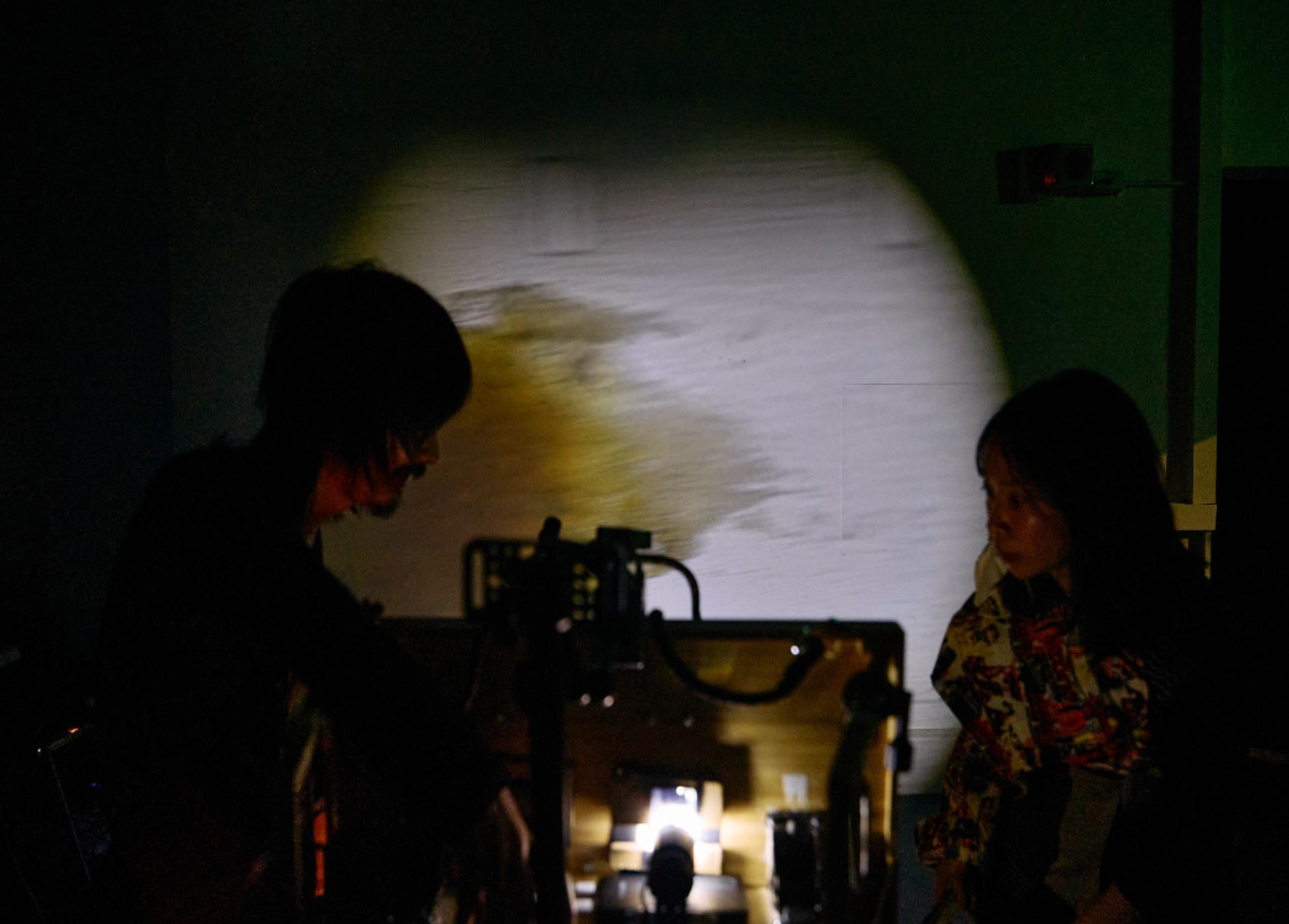 Two performers work in near darkness, illuminated by a projector casting light on the wall between them.