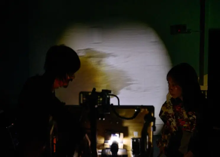 Two performers work in near darkness, illuminated by a projector casting light on the wall between them.