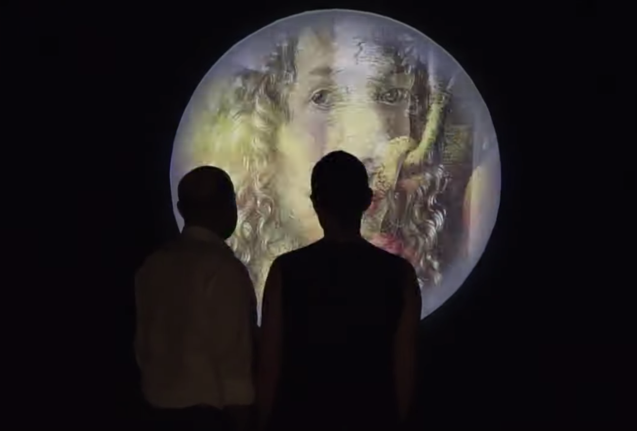 Two individuals are seen from behind, gazing at a projection of an old painted portrait with long curly hair, displayed on...