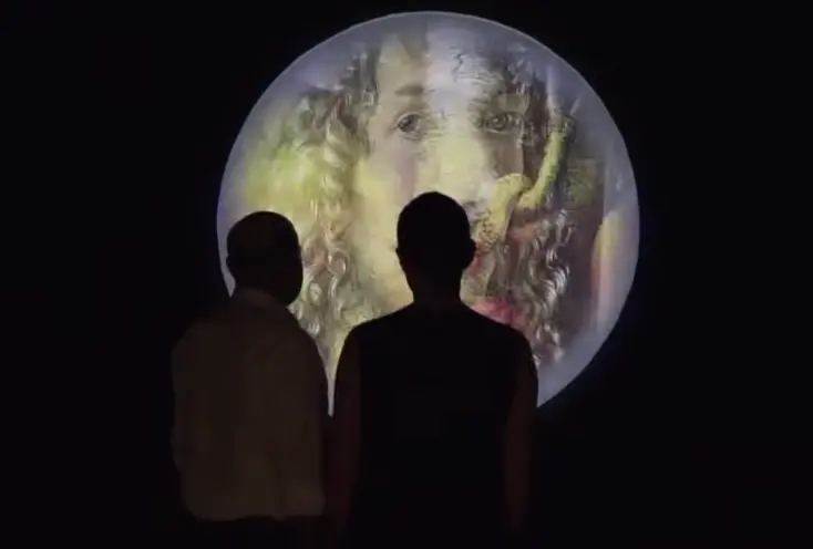 Two individuals are seen from behind, gazing at a projection of an old painted portrait with long curly hair, displayed on...