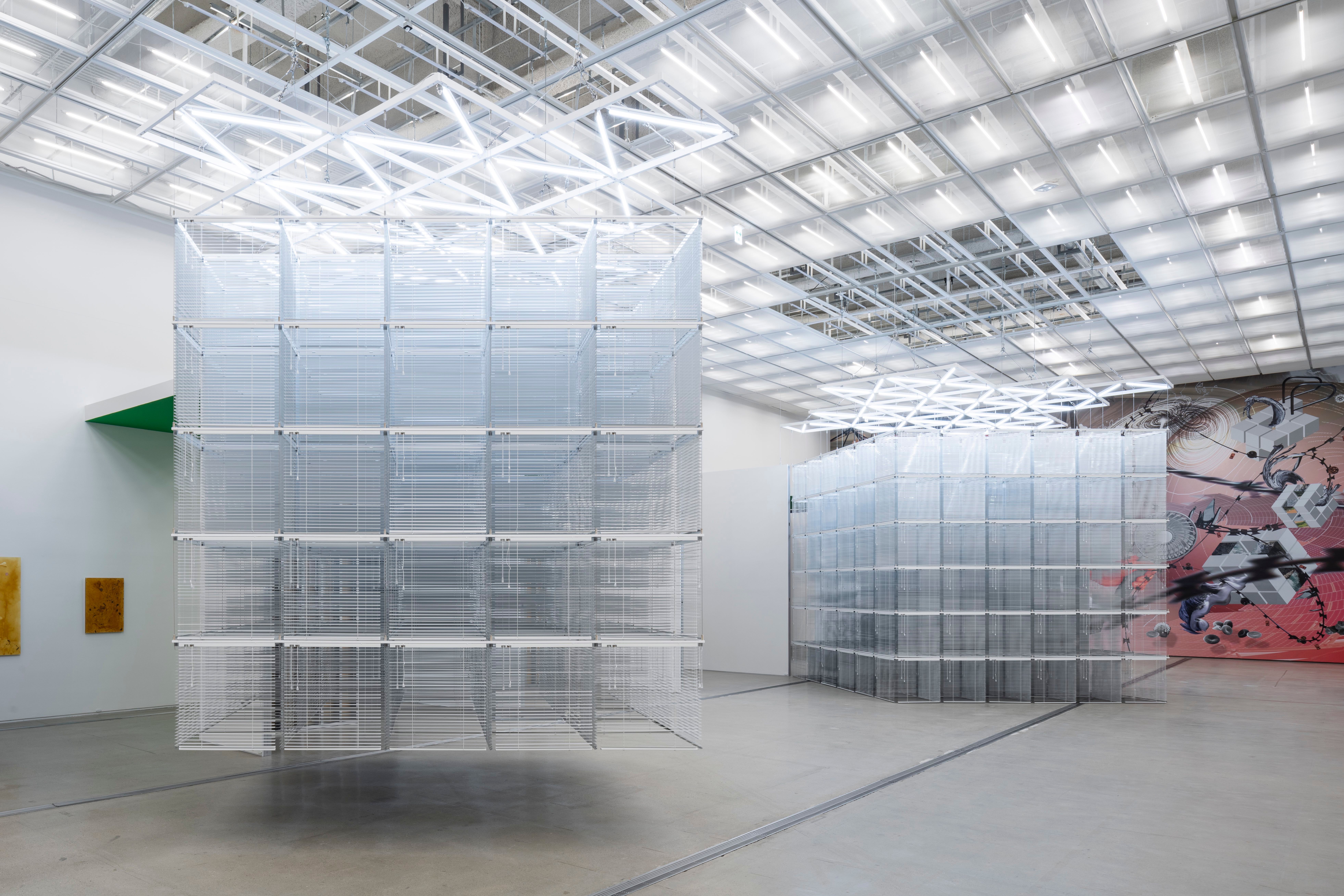 Two transparent cubic sculptures, one suspended from the ceiling and one on the floor, in a brightly lit art exhibition ro...