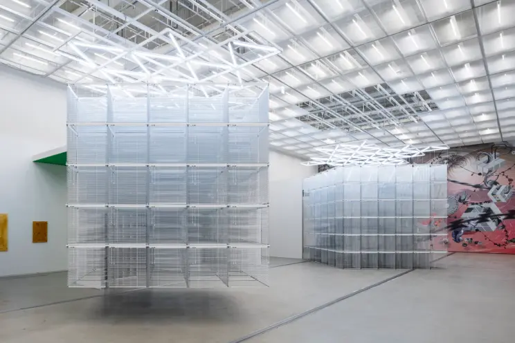 Two transparent cubic sculptures, one suspended from the ceiling and one on the floor, in a brightly lit art exhibition ro...