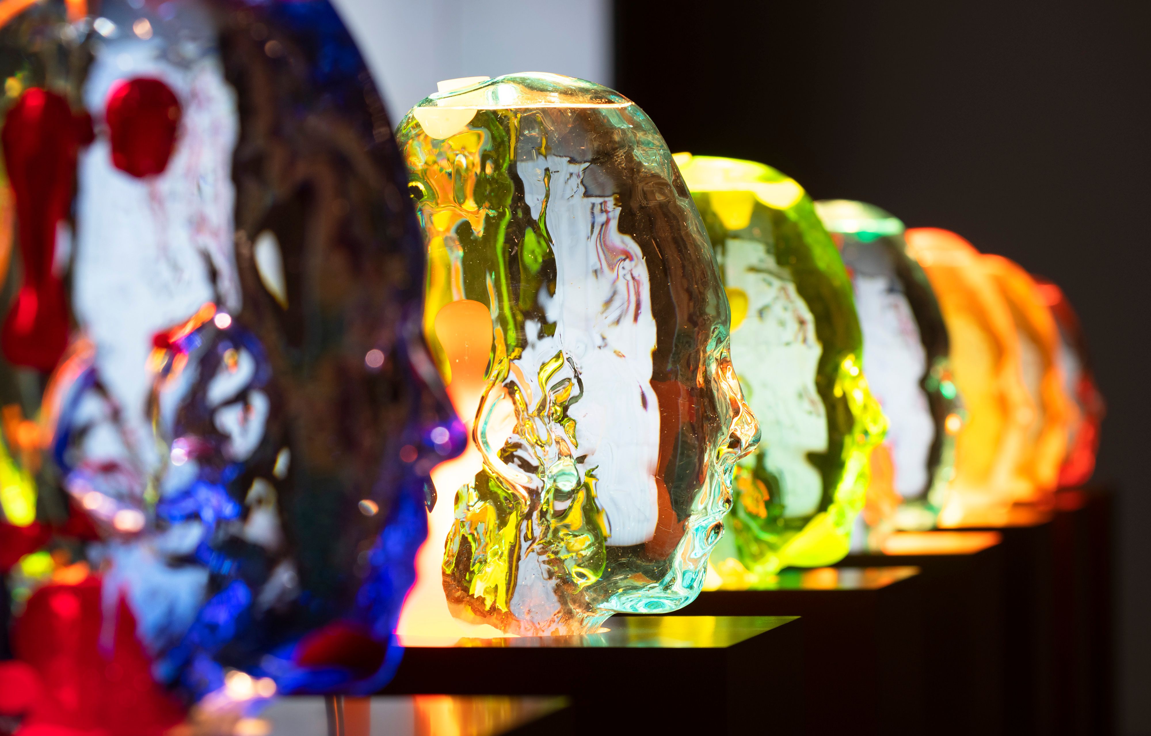 A row of brightly colored, translucent glass head sculptures glowing under light.