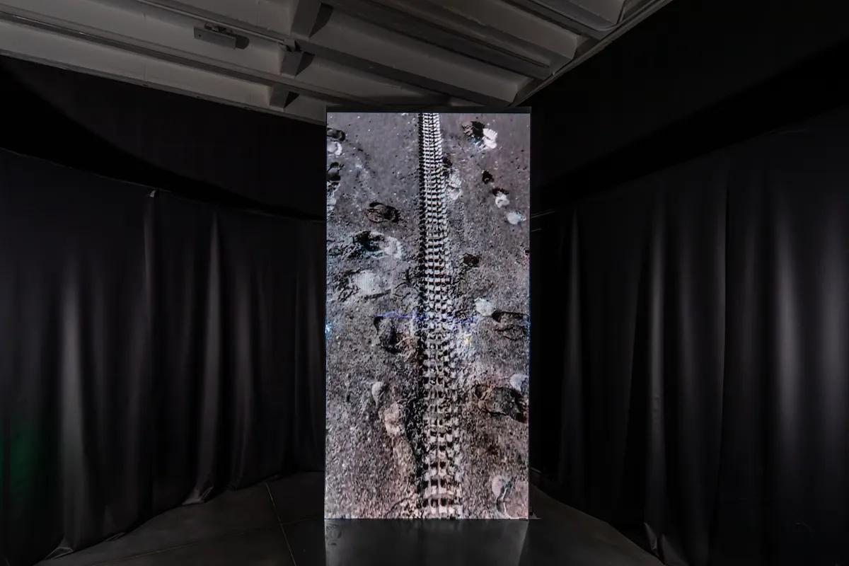 An art installation featuring a dark floor imprinted with footprints and a tire track, creating an aesthetic reminiscent o...