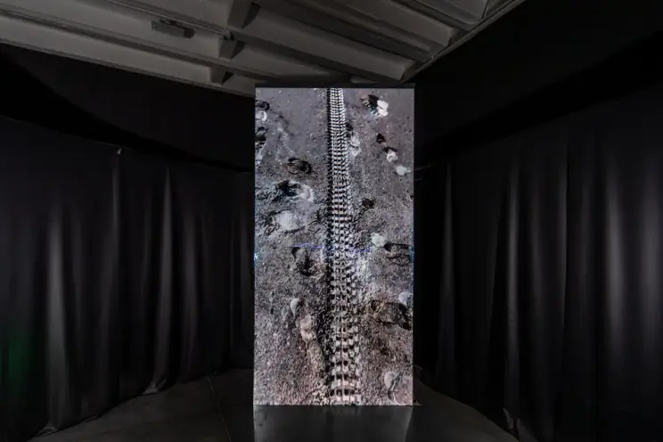 An art installation featuring a dark floor imprinted with footprints and a tire track, creating an aesthetic reminiscent o...