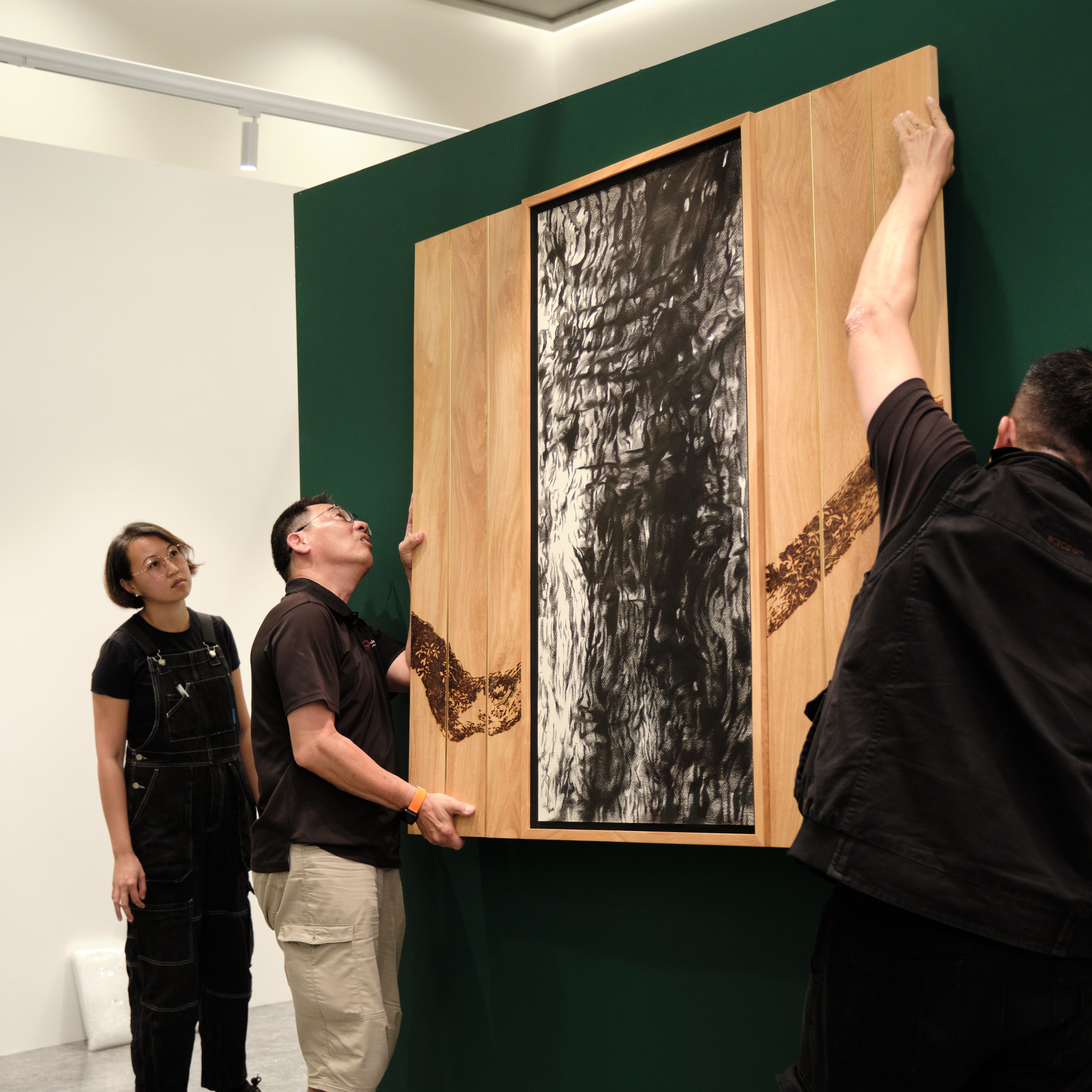 Three people install or examine a large framed artwork with a charcoal drawing and wood panels featuring a burnt branch design.