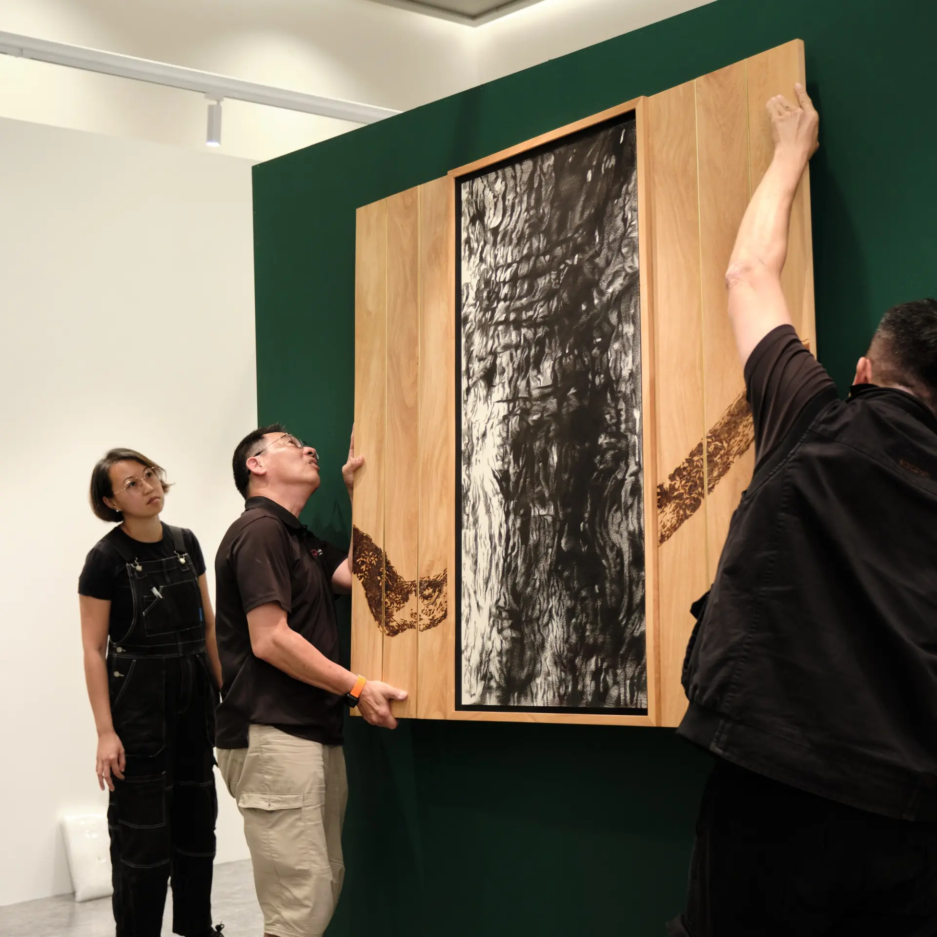 Three people install or examine a large framed artwork with a charcoal drawing and wood panels featuring a burnt branch design.